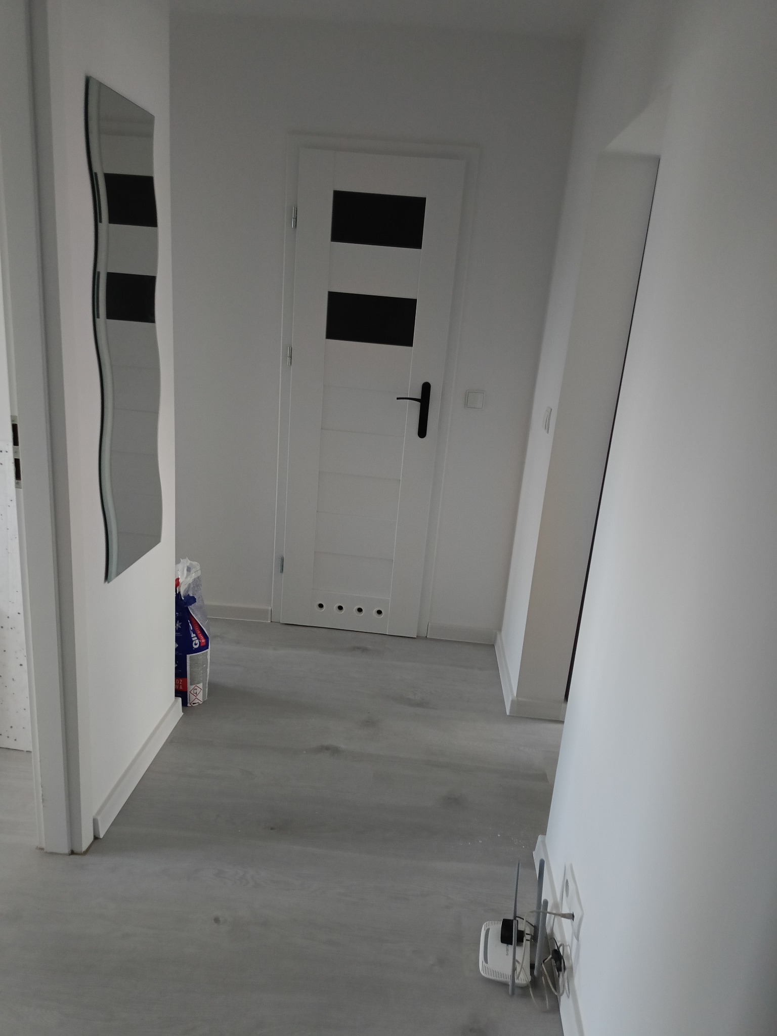 Wnętrze przedpokoju z szarą podłogą, białą ścianą i drzwiami z czarnymi poziomymi paskami, widoczne lustro o nieregularnym kształcie i wystający worek z materiałami budowlanymi.