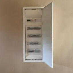 Otwarta, biała skrzynka rozdzielcza z bezpiecznikami na beżowej ścianie, widoczne bezpieczniki i oznaczenia numerów obwodów.