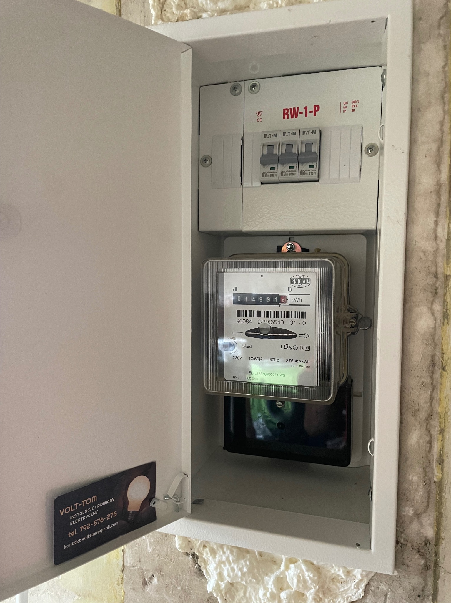 Otwarta skrzynka elektryczna z licznikiem energii i wyłącznikami, widoczna wizytówka firmy elektrycznej Volt-Tom przymocowana do drzwiczek, ściana z pianką montażową w tle.