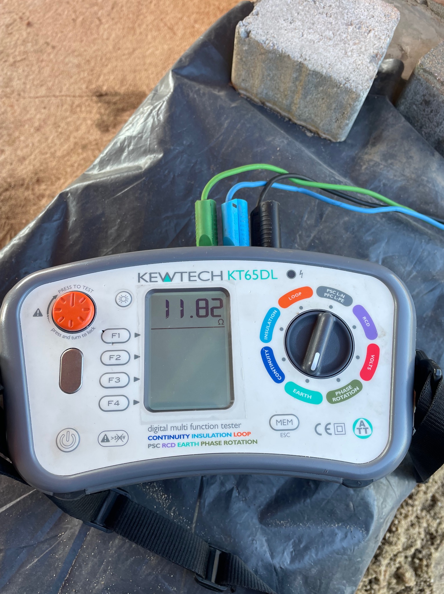 Miernik wielofunkcyjny Kewtech KT65DL z podłączonymi przewodami pomiarowymi, leżący na czarnej folii, obok betonowych kostek. Wyświetlacz pokazuje wartość 11.82 omów.