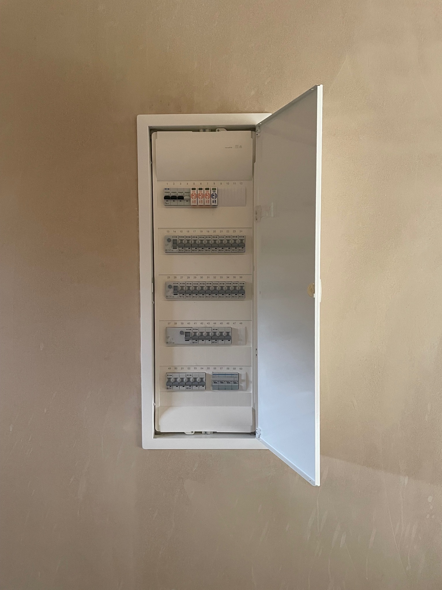 Otwarta, biała skrzynka rozdzielcza z bezpiecznikami na beżowej ścianie, widoczne bezpieczniki i oznaczenia numerów obwodów.