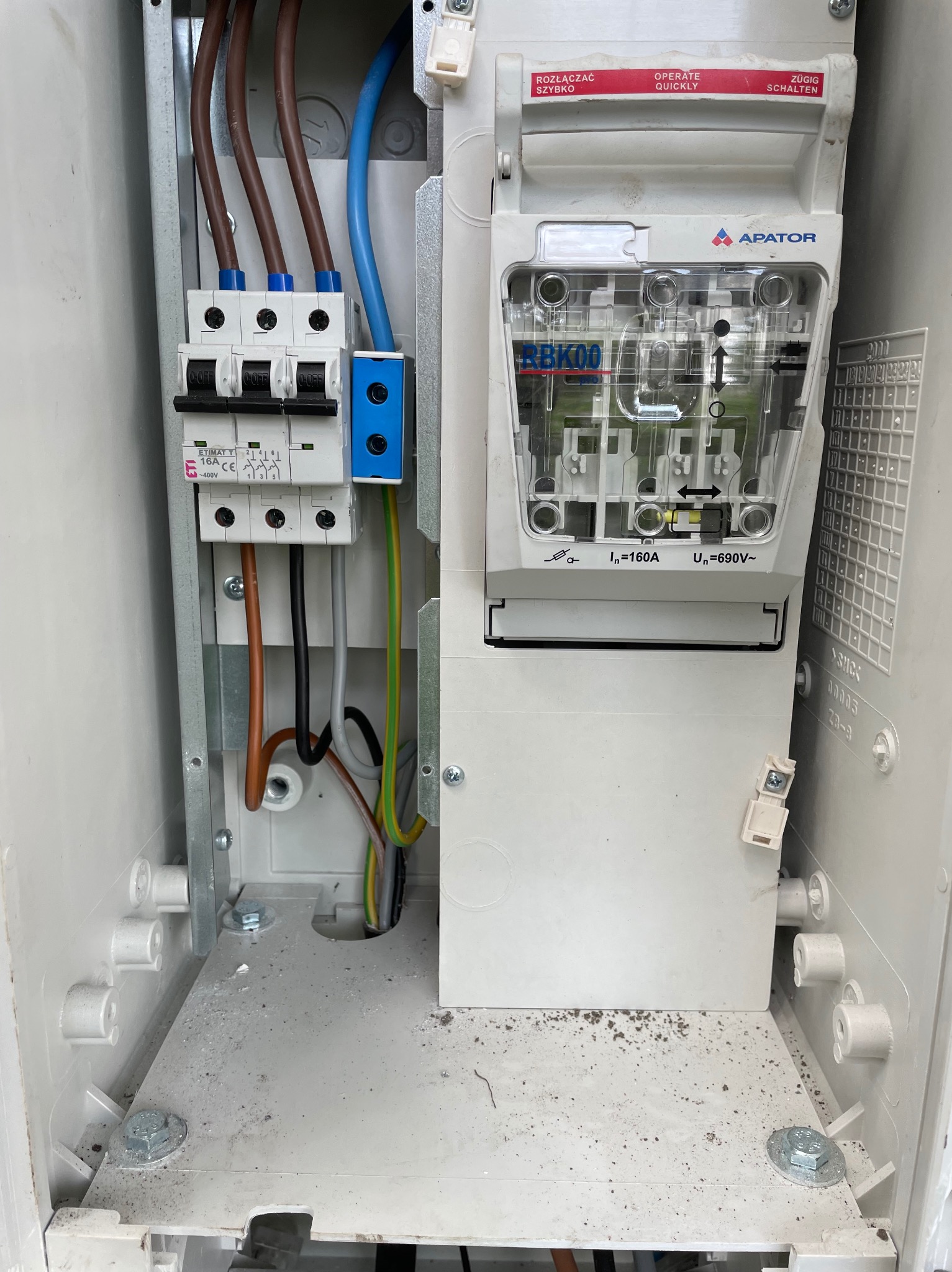 Wnętrze skrzynki elektrycznej z zainstalowanymi bezpiecznikami i wyłącznikiem, widoczne przewody w różnych kolorach, elementy montażowe i oznaczenia producenta Apator.