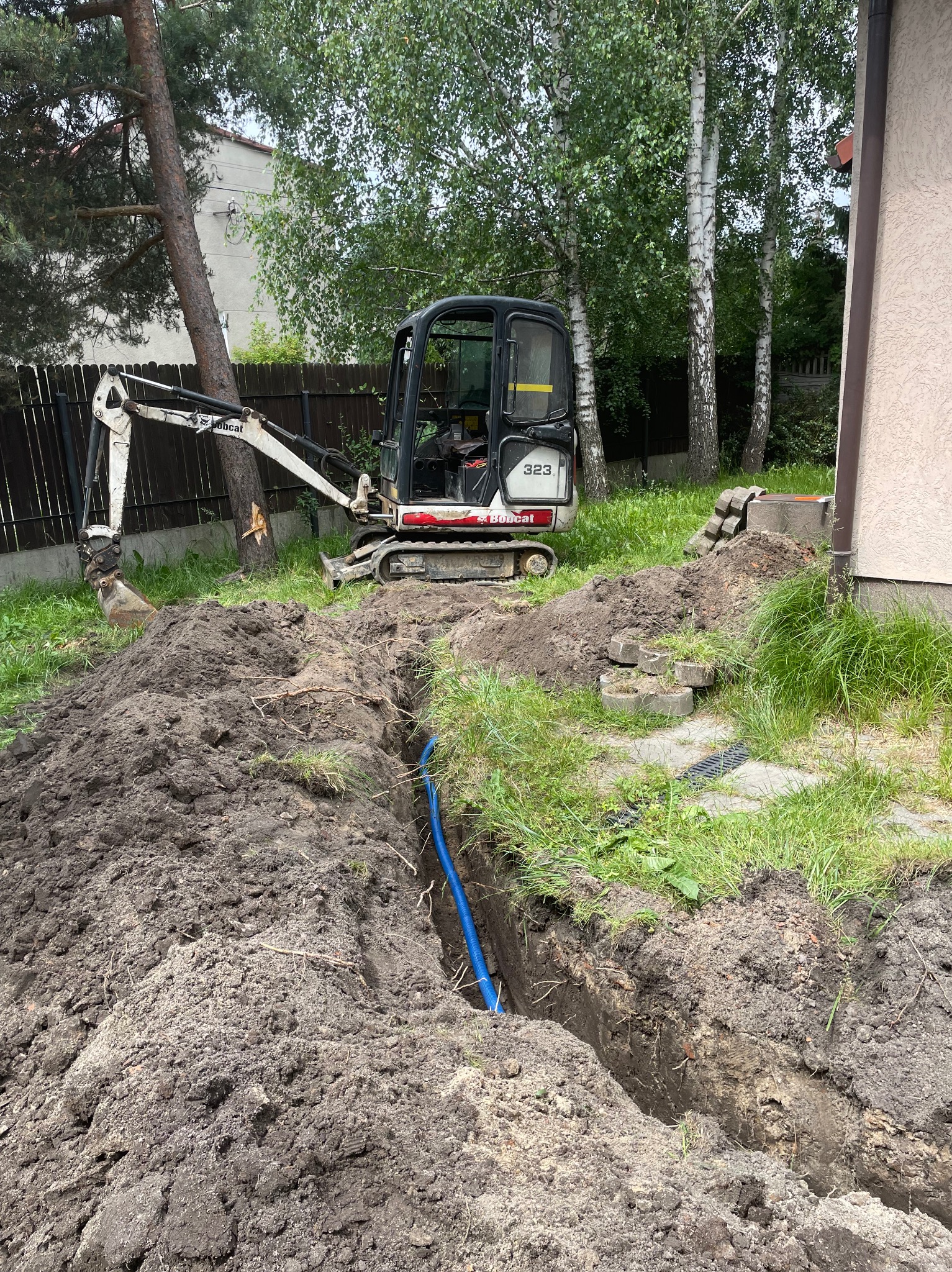 Mini koparka Bobcat 323 obok wykopanego rowu z niebieską rurą w ziemi, przygotowanie do ułożenia kabla elektrycznego w gruncie.
