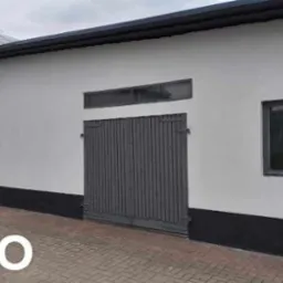 Biały budynek gospodarczy z szarą, drewnianą bramą, małym oknem i czarną podmurówką, widziany z poziomu brukowanego podjazdu.