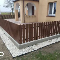 Nowo postawione, brązowe, drewniane ogrodzenie z betonową podmurówką, otaczające taras domu jednorodzinnego z beżową elewacją. Białe kamienie dekoracyjne u podstawy ogrodzenia.