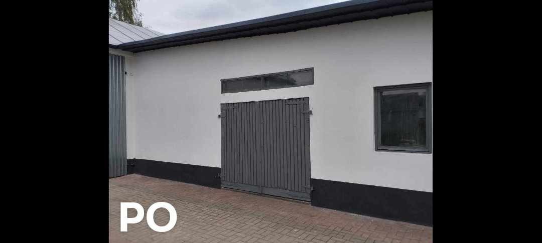 Biały budynek gospodarczy z szarą, drewnianą bramą, małym oknem i czarną podmurówką, widziany z poziomu brukowanego podjazdu.