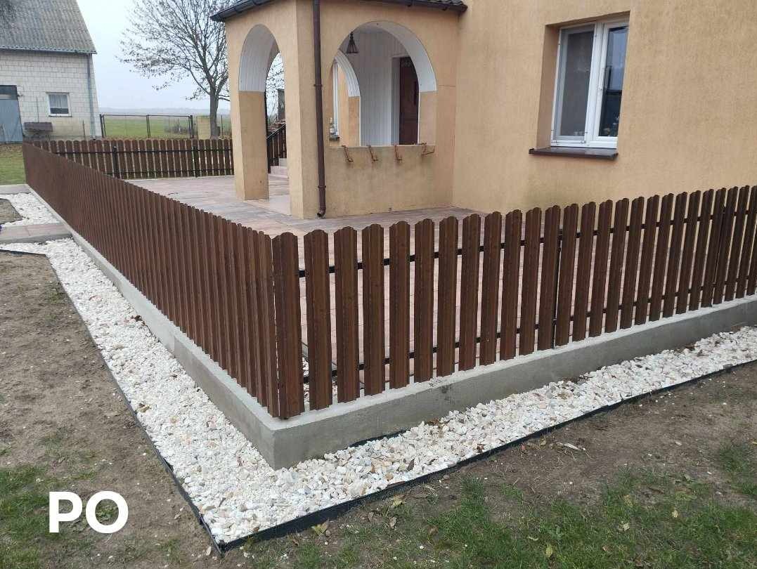 Nowo postawione, brązowe, drewniane ogrodzenie z betonową podmurówką, otaczające taras domu jednorodzinnego z beżową elewacją. Białe kamienie dekoracyjne u podstawy ogrodzenia.