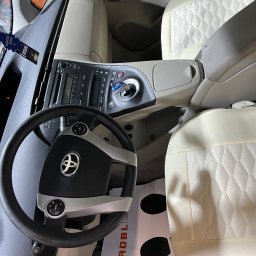 CAR MAX - Wnętrze samochodu z jasną tapicerką foteli z przeszyciami w romby, widoczna kierownica z logo Toyota, deska rozdzielcza oraz zapach samochodowy Wunder-Baum New Car.