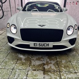 NOVA Detailing. Myjnia ręczna, korekta lakieru, pranie tapicerki - Biały Bentley Continental GT z włączonymi światłami, uchwycony od przodu w myjni samochodowej, na mokrej posadzce z resztkami piany i wężami na szarej ścianie.