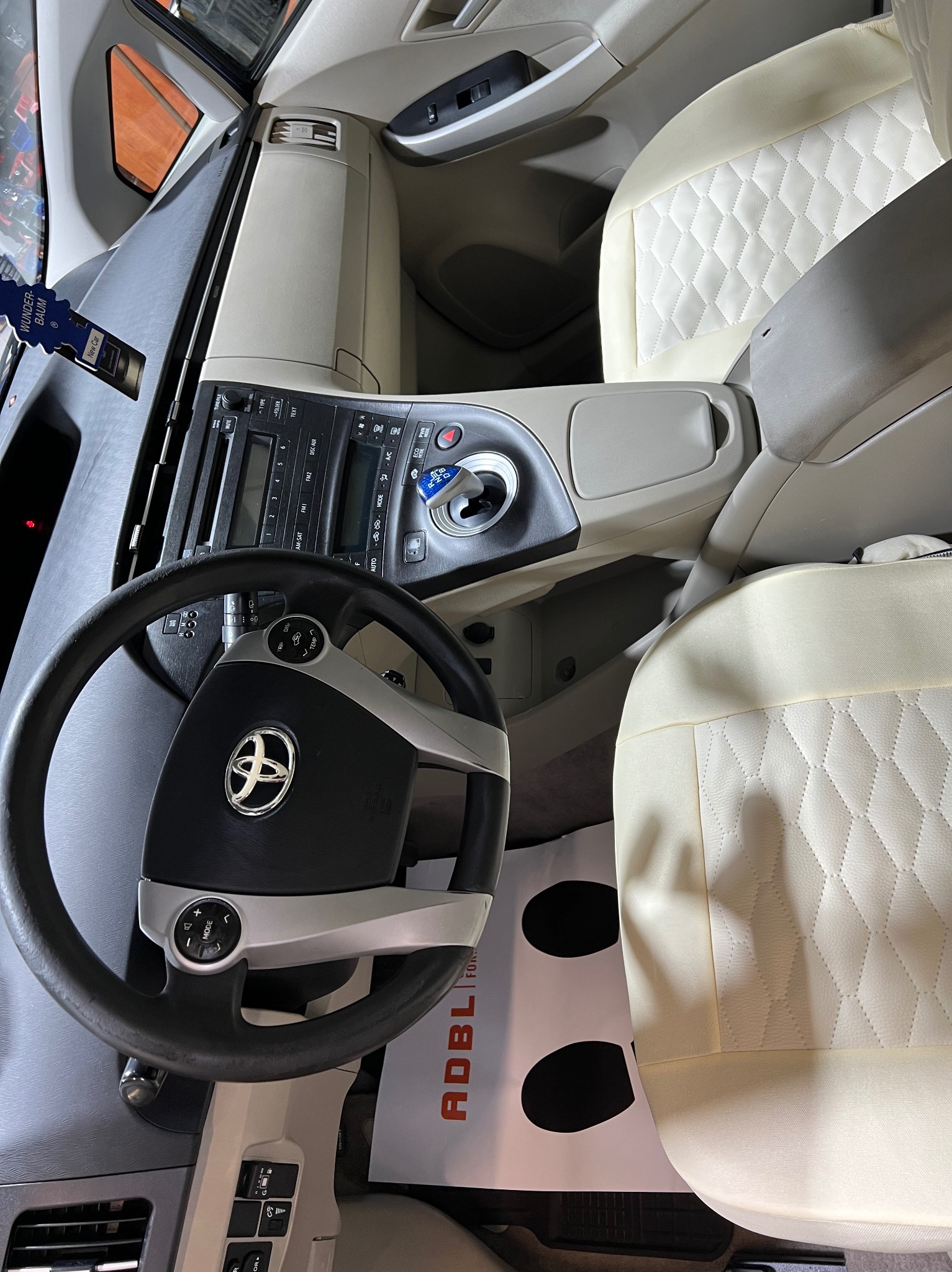 Wnętrze samochodu z jasną tapicerką foteli z przeszyciami w romby, widoczna kierownica z logo Toyota, deska rozdzielcza oraz zapach samochodowy Wunder-Baum New Car.