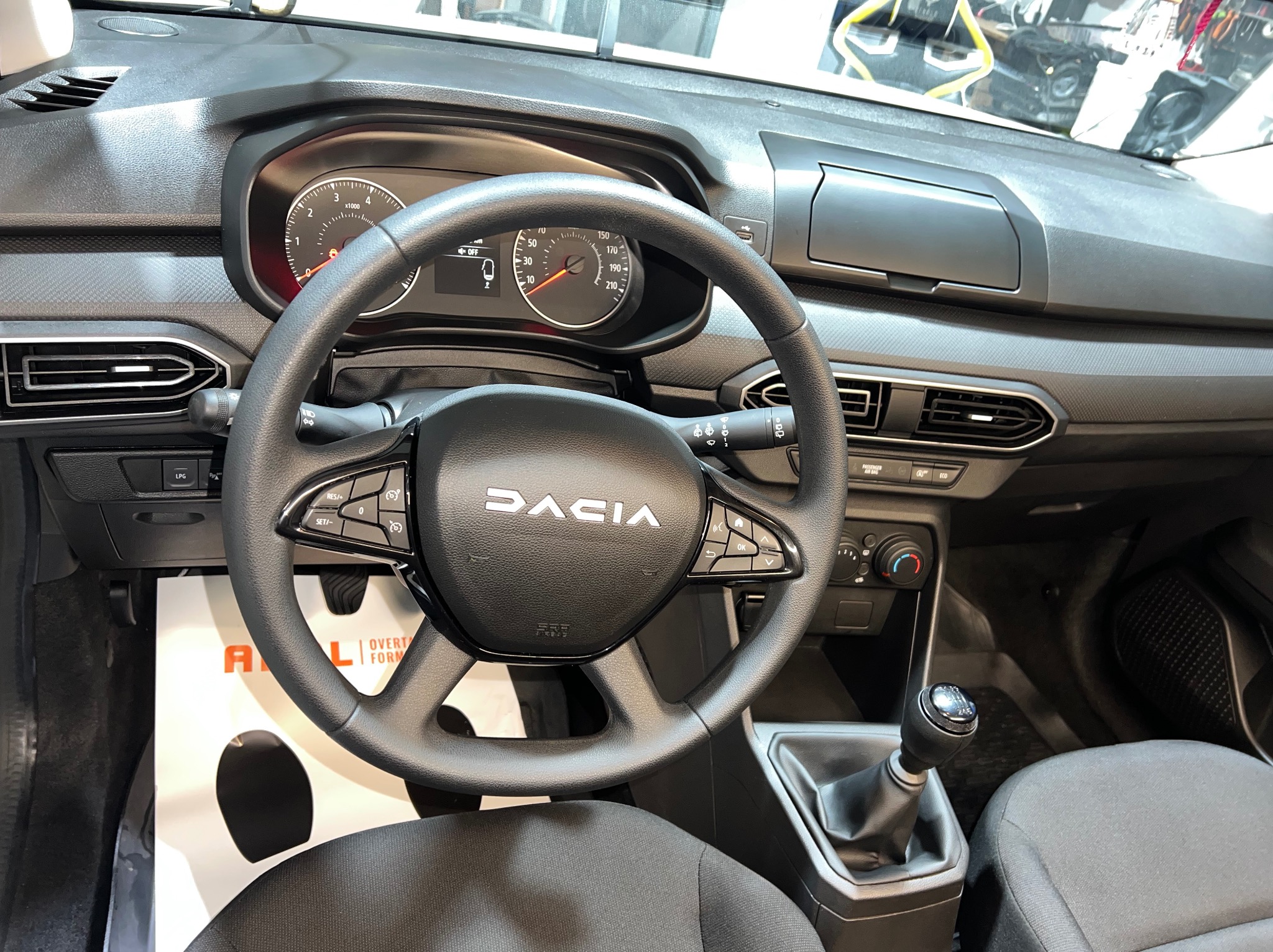 Wnętrze samochodu Dacia z widoczną deską rozdzielczą, kierownicą z logo i dźwignią zmiany biegów. Widoczny licznik prędkości i obrotomierz.