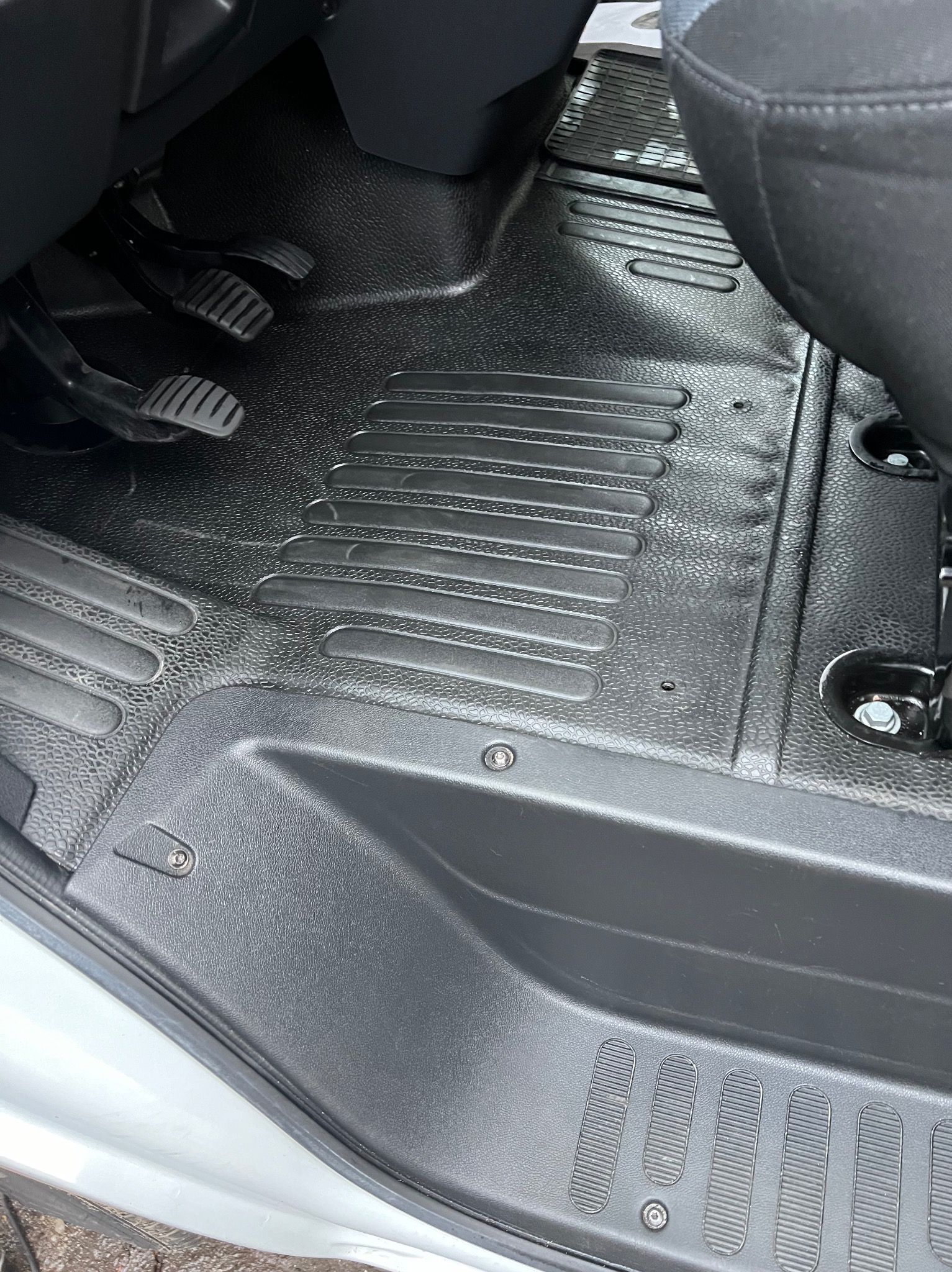 Czarne, gumowe dywaniki z wzorem w podłodze samochodu dostawczego, widoczne pedały i mocowania fotela.