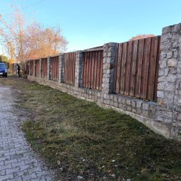 GRACO GRACJAN KOZIEŁ - Długi, drewniano-kamienny płot oddzielający chodnik od posesji. Świeżo skoszona trawa przy płocie. W tle zaparkowany niebieski bus i osoba pracująca przy płocie.