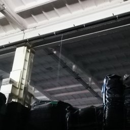 GRACO GRACJAN KOZIEŁ - Wnętrze hali magazynowej z betonowym stropem kasetonowym, widoczne czarne worki z zawartością, oświetlenie przemysłowe i fragmenty konstrukcji budynku.