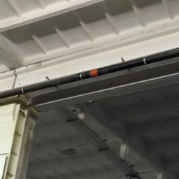 GRACO GRACJAN KOZIEŁ - Czarne rury instalacji pod stropem w hali przemysłowej, wsparte na metalowej konstrukcji. Widoczne elementy mocujące i fragment pomarańczowej taśmy na rurze.