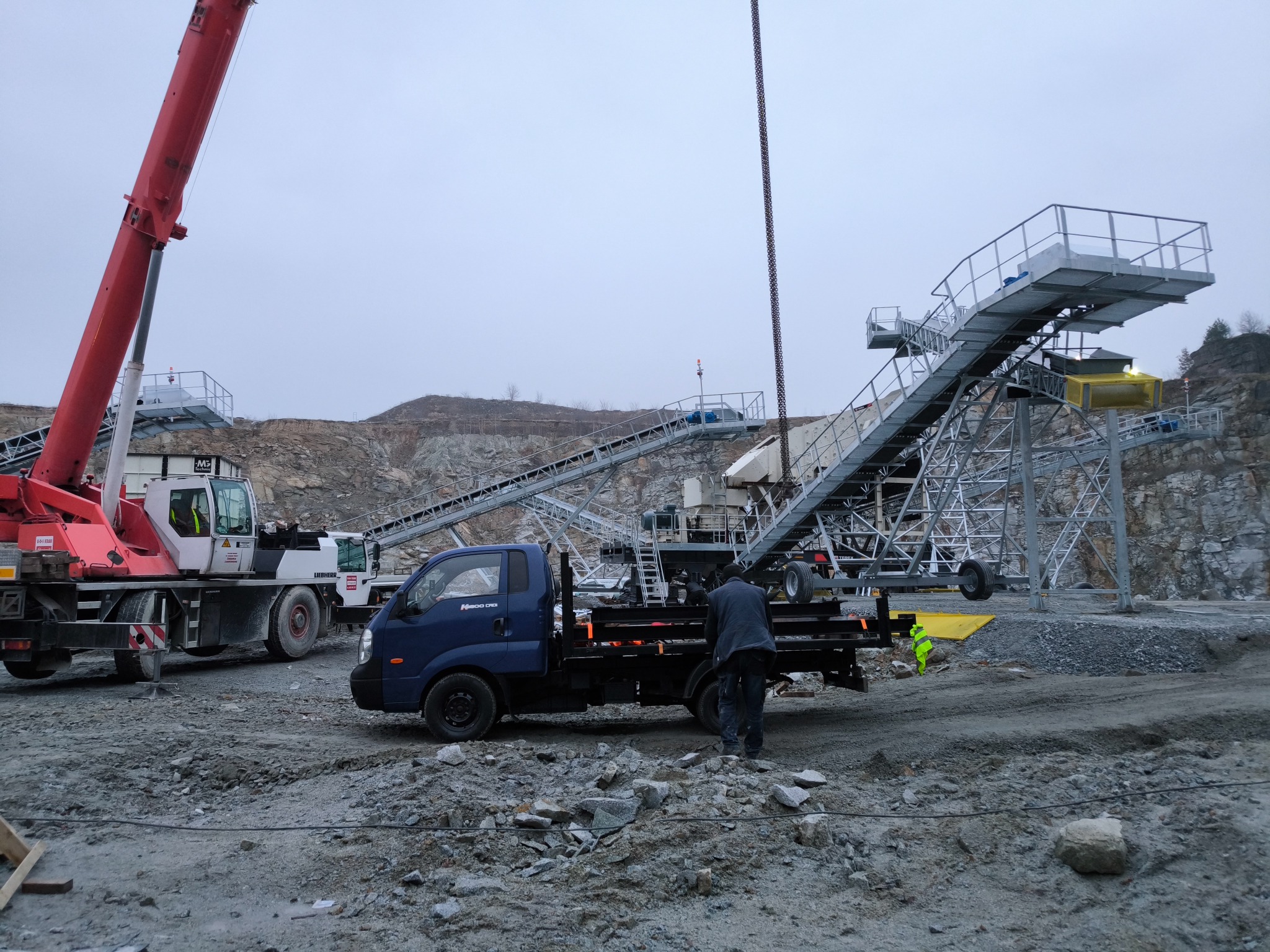 Montaż linii technologicznej w kamieniołomie. Żuraw i ciężarówka w trakcie pracy. Widoczny pracownik w roboczym ubraniu. Szaro-błękitne niebo.