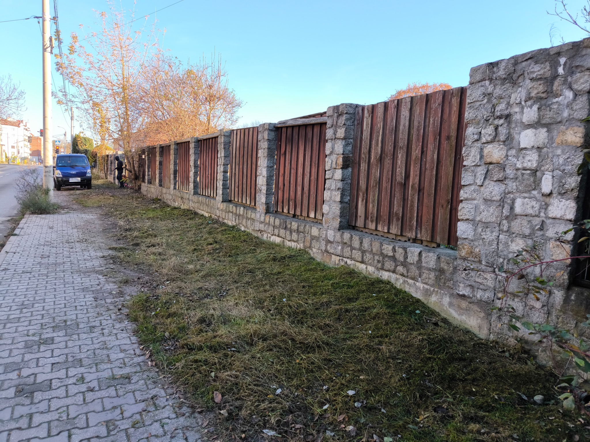 Długi, drewniano-kamienny płot oddzielający chodnik od posesji. Świeżo skoszona trawa przy płocie. W tle zaparkowany niebieski bus i osoba pracująca przy płocie.