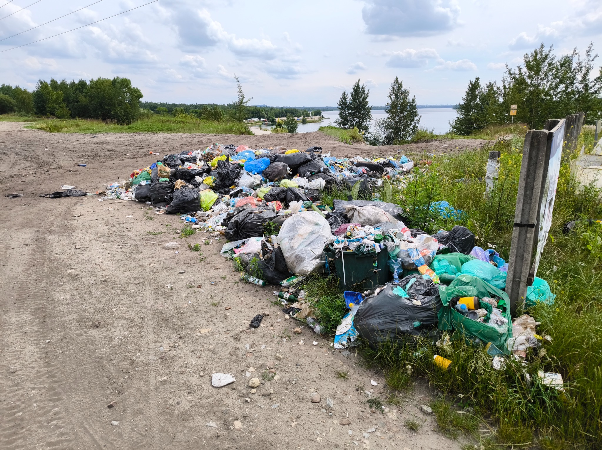 Nielegalne składowisko odpadów z workami i śmieciami na poboczu drogi gruntowej, w tle jezioro i drzewa. Widok z poziomu gruntu, pochmurne niebo.