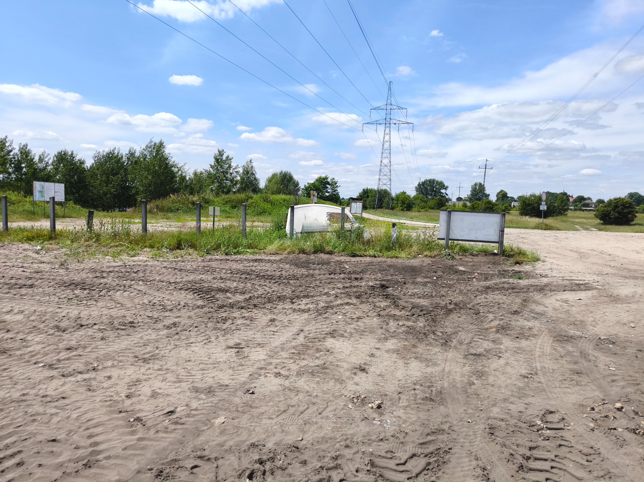Teren po robotach ziemnych z widocznymi śladami opon, słupami energetycznymi w tle i fragmentami zieleni. Błękitne niebo z chmurami w słoneczny dzień.