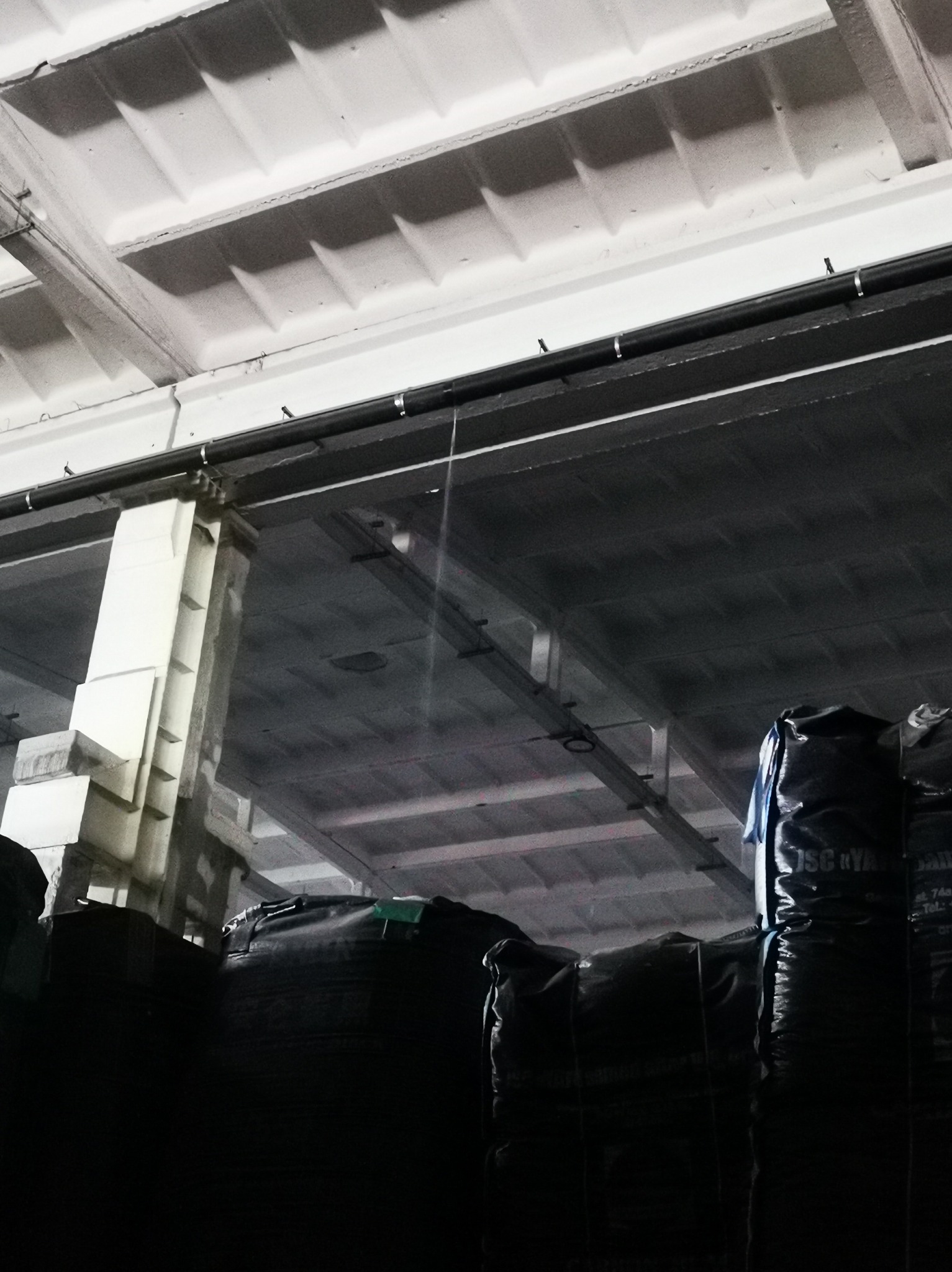 Wnętrze hali magazynowej z betonowym stropem kasetonowym, widoczne czarne worki z zawartością, oświetlenie przemysłowe i fragmenty konstrukcji budynku.