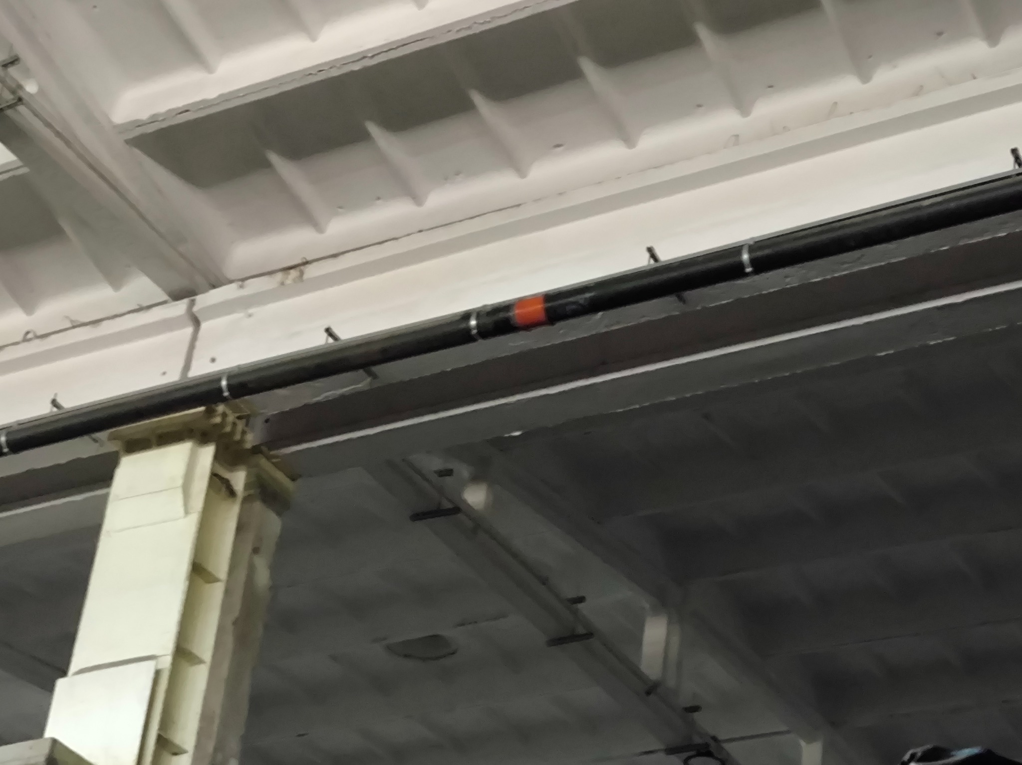 Czarne rury instalacji pod stropem w hali przemysłowej, wsparte na metalowej konstrukcji. Widoczne elementy mocujące i fragment pomarańczowej taśmy na rurze.