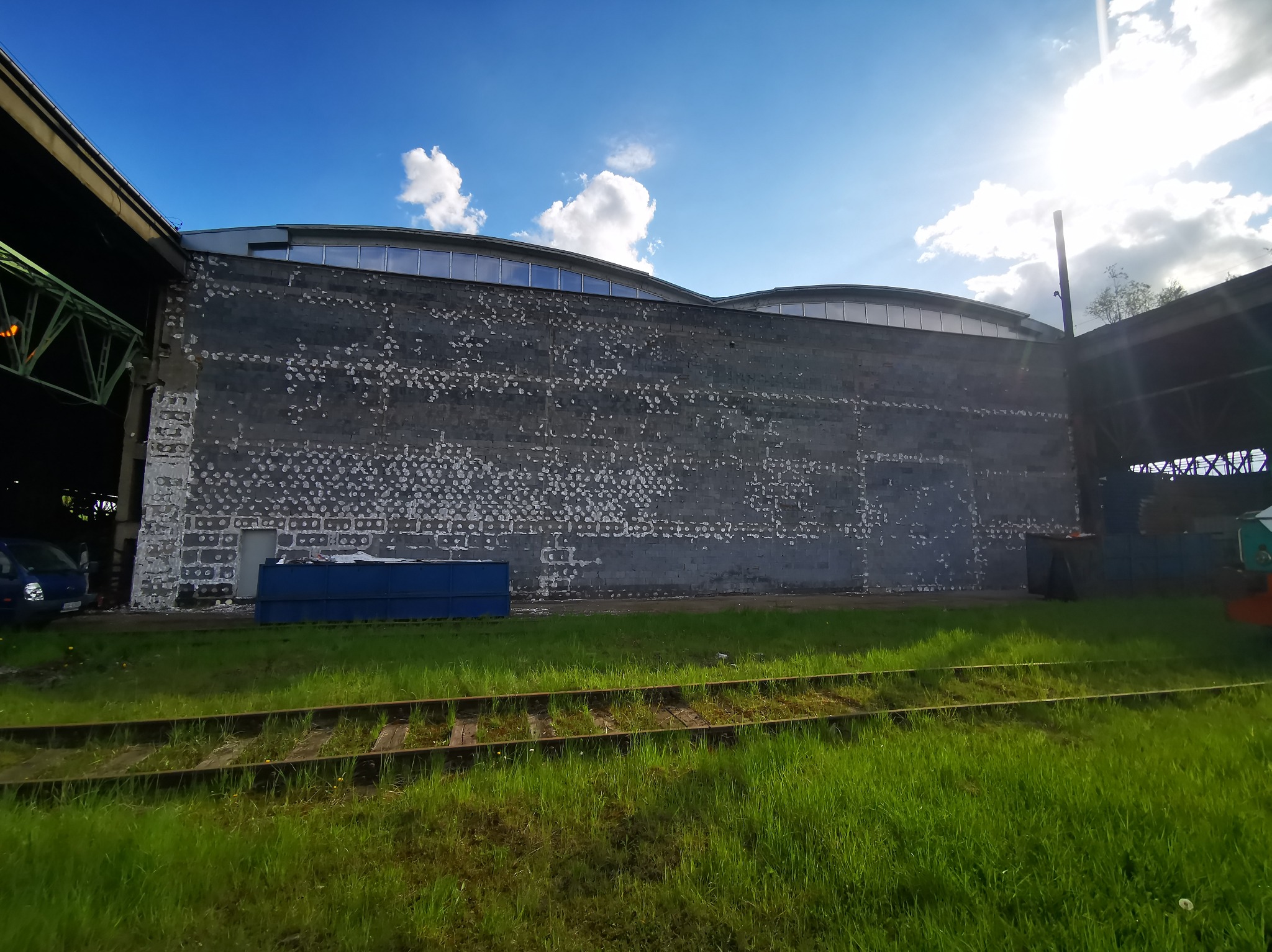 Szara ściana budynku z odpadającą farbą, niebieski kontener, fragment torów kolejowych i trawa na pierwszym planie, błękitne niebo z chmurami w tle.