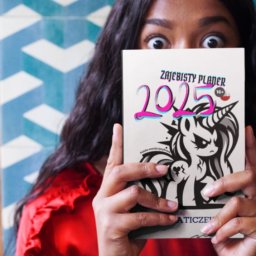 Patrycja Ciborowska - Kobieta w Katowicach, ubrana w czerwoną bluzkę, trzyma przed twarzą planer 'Zajebisty Planer 2025' z ilustracją złego jednorożca.