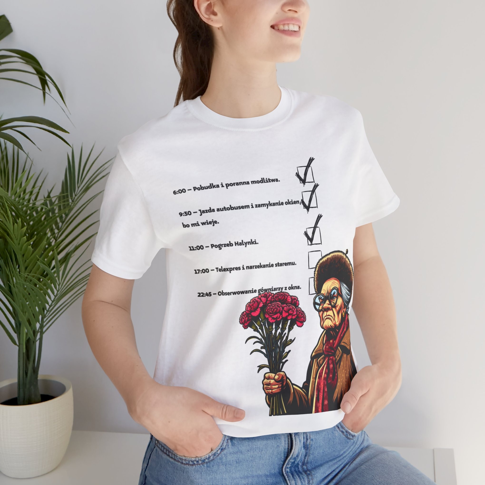 Kobieta w dżinsach pozuje w białym t-shircie z nadrukiem humorystycznej listy zadań i postaci starszej pani z bukietem kwiatów.