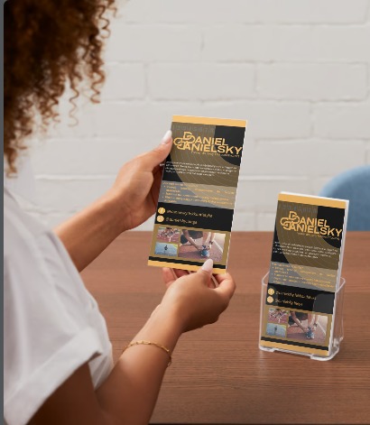 Kobieta trzyma ulotkę z logo i informacjami o Daniel Danielsky, obok stojak z ulotkami na drewnianym stole.
