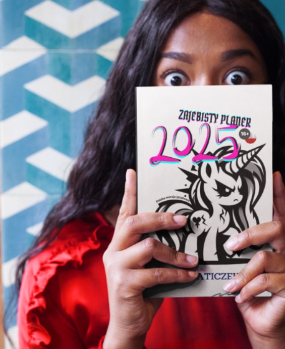 Kobieta w Katowicach, ubrana w czerwoną bluzkę, trzyma przed twarzą planer 'Zajebisty Planer 2025' z ilustracją złego jednorożca.