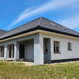 Nowoczesny dom parterowy z szarą dachówką, jasnoszarą elewacją i drewnianymi oknami, widok z zewnątrz w słoneczny dzień.