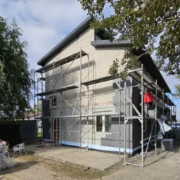 Dwukondygnacyjny dom w trakcie ocieplania elewacji, widoczne rusztowania, częściowo ocieplone ściany styropianem grafitowym, worki z zaprawą na ziemi.