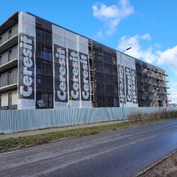 BESTHOUSE4YOU SPÓŁKA Z OGRANICZONĄ ODPOWIEDZIALNOŚCIĄ - Budynek wielorodzinny w trakcie prac elewacyjnych, osłonięty siatką z logo Cerersit, widoczne rusztowania i fragment ulicy.
