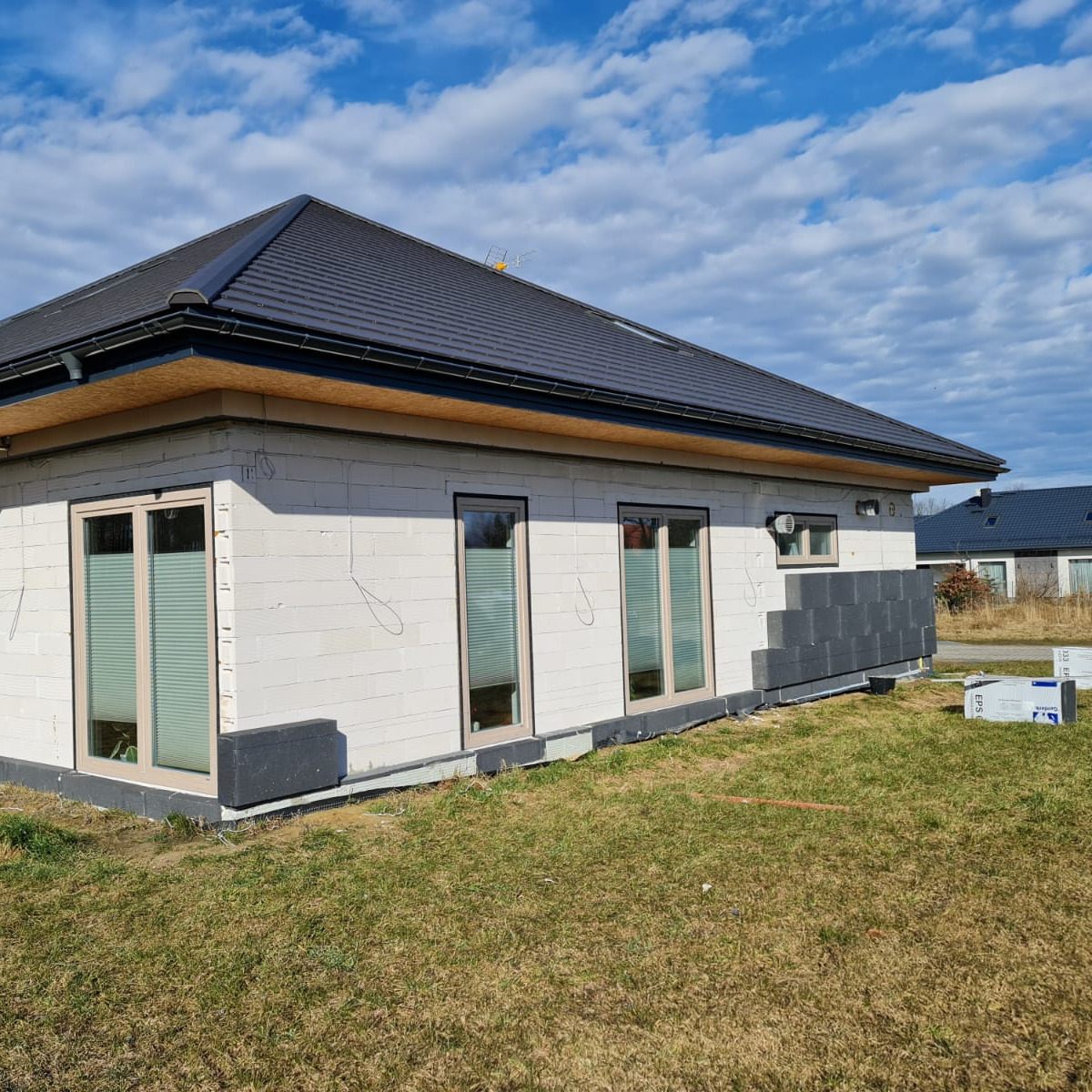 Niedokończony dom jednorodzinny z białego bloczka, z oknami z żaluzjami wewnętrznymi, częściowo obłożony szarymi płytami izolacyjnymi, widoczne płyty styropianowe na trawie, błękitne niebo...