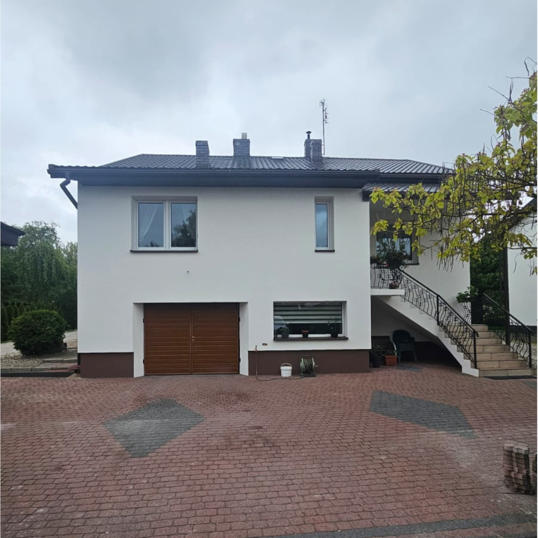 Dwupiętrowy dom z białymi ścianami i brązowym akcentem u dołu, brązowa brama garażowa i schody z kutą balustradą prowadzące na balkon, widoczne rośliny w doniczkach na balkonie, kostka brukowa...