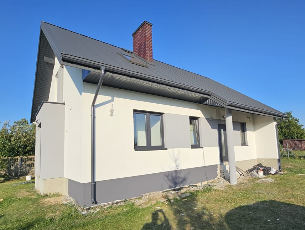 Nowoczesny dom jednorodzinny z jasną elewacją, szarą podmurówką i grafitowym dachem z ceglanym kominem, widok z niskiej perspektywy.