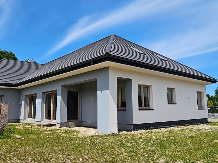 Nowoczesny dom parterowy z szarą dachówką, jasnoszarą elewacją i drewnianymi oknami, widok z zewnątrz w słoneczny dzień.