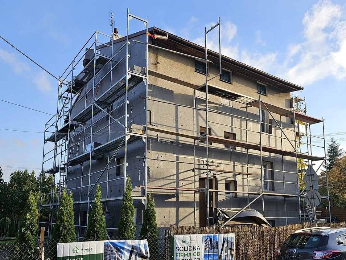 Dwukondygnacyjny dom w trakcie prac, osłonięty rusztowaniem i pokryty szarymi płytami izolacyjnymi; widoczny pracownik na górze rusztowania.