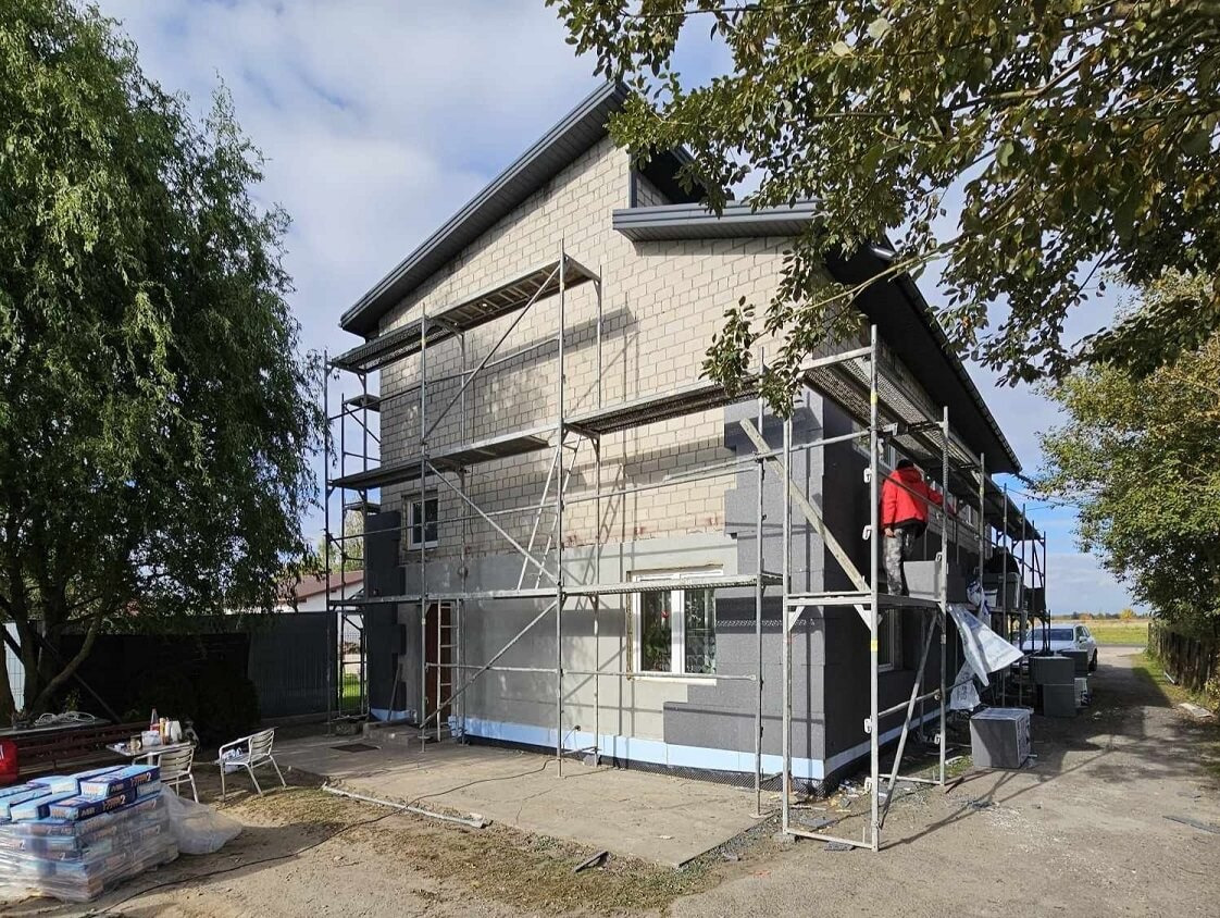 Dwukondygnacyjny dom w trakcie ocieplania elewacji, widoczne rusztowania, częściowo ocieplone ściany styropianem grafitowym, worki z zaprawą na ziemi.
