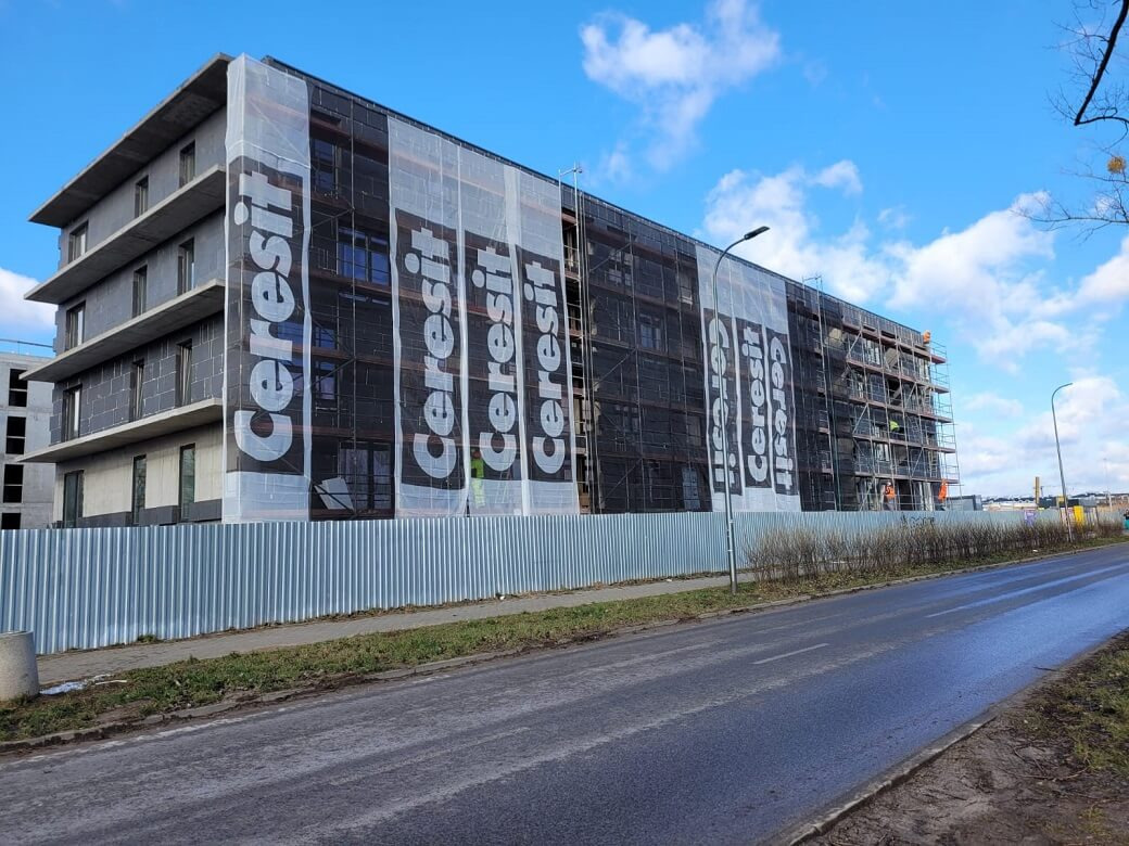 Budynek wielorodzinny w trakcie prac elewacyjnych, osłonięty siatką z logo Cerersit, widoczne rusztowania i fragment ulicy.