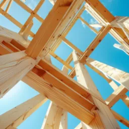 Drewniana konstrukcja dachu domu w trakcie budowy, widok od dołu na tle błękitnego nieba.