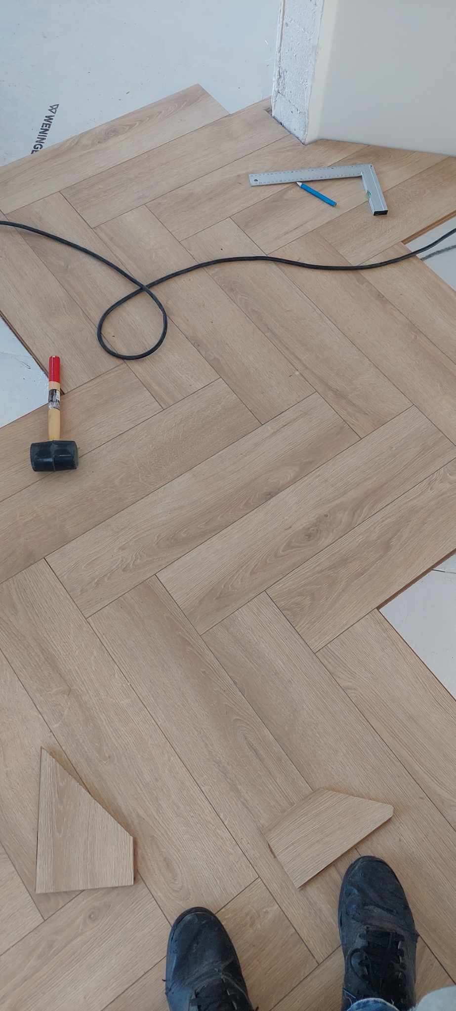 Ułożone panele podłogowe w jodełkę, widoczny gumowy młotek, kątownik, ołówek i docięty element panela oraz fragment obuwia osoby wykonującej prace.