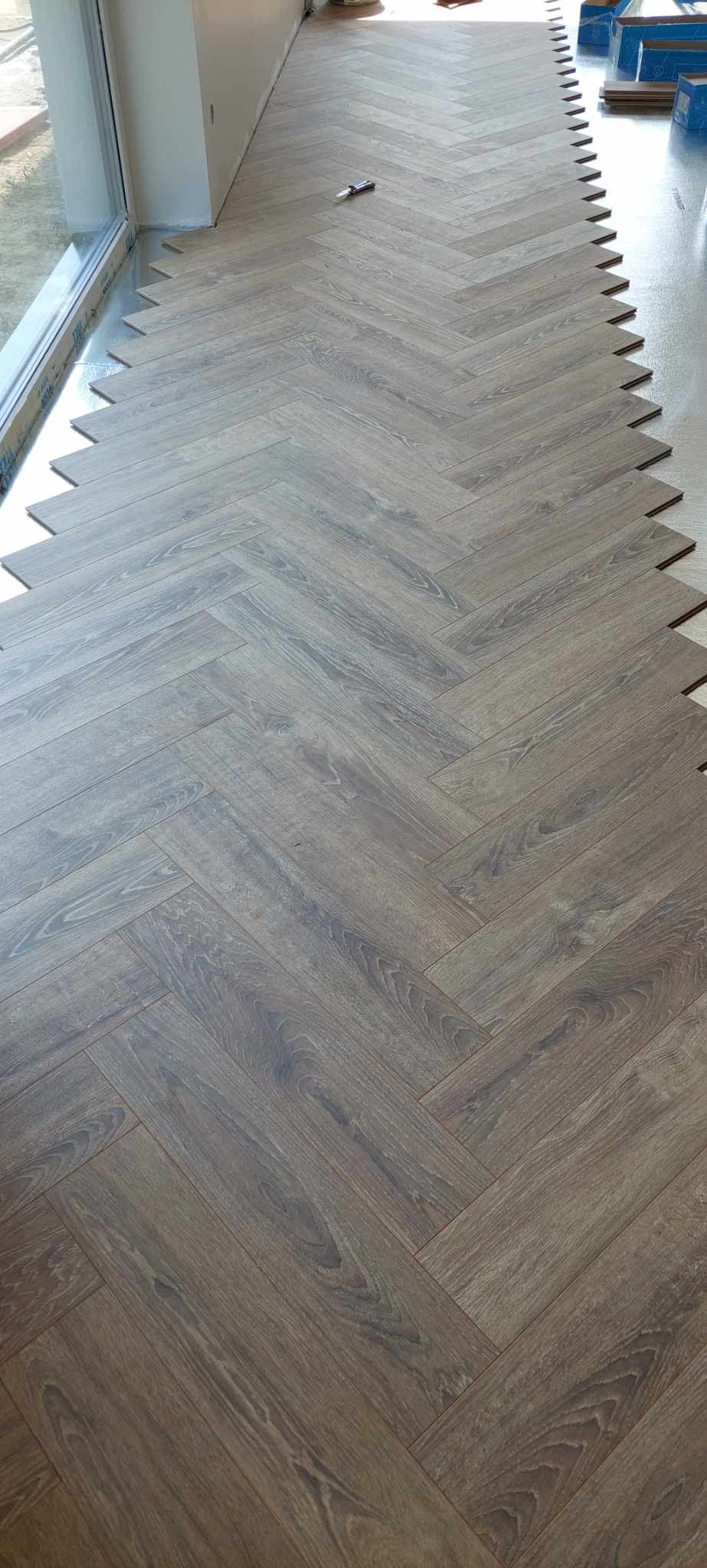 Parkiet ułożony we wzór jodełki, przycięty przy ścianie, z widocznymi elementami montażowymi i narzędziem na podłodze.