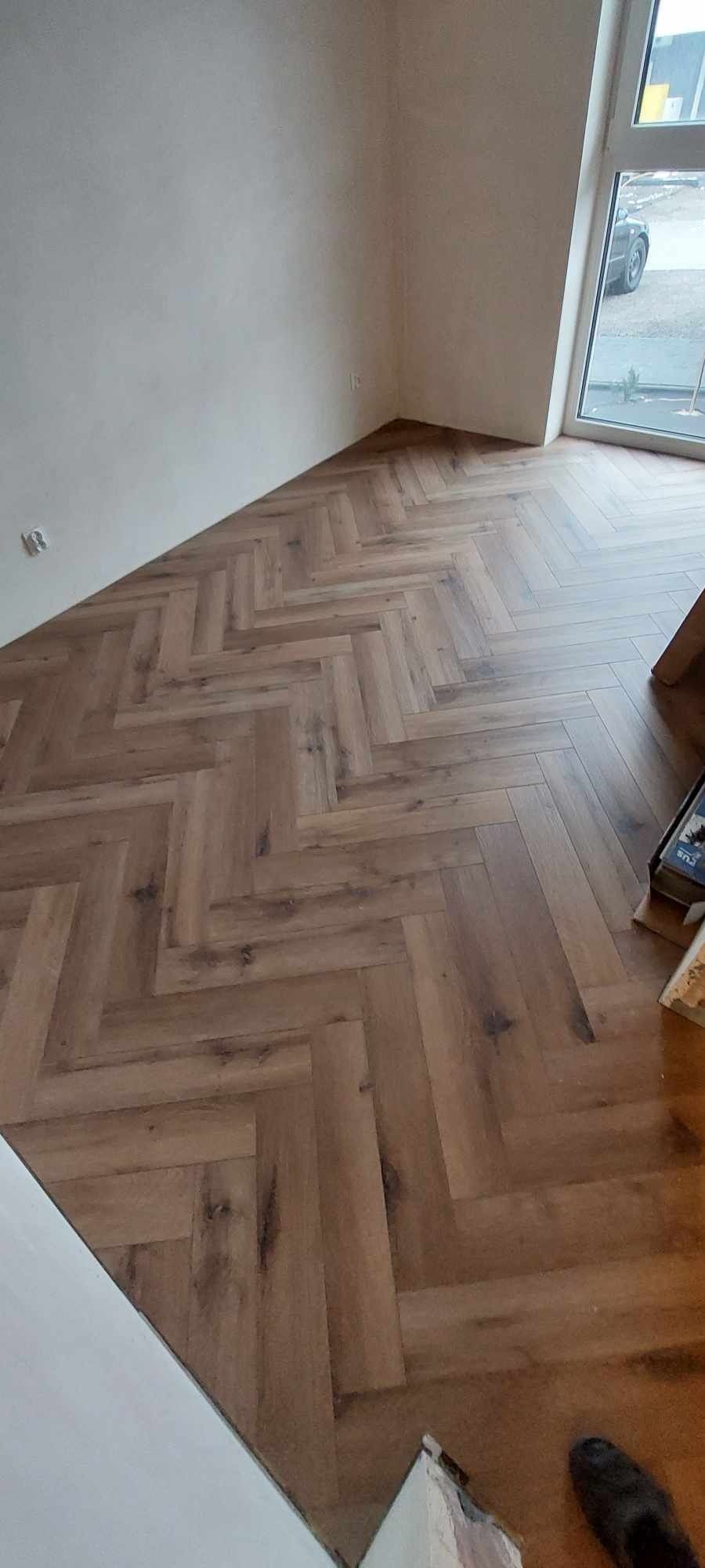 Ułożona podłoga z paneli w jodełkę, widok na jasne ściany i okno z widokiem na ulicę.