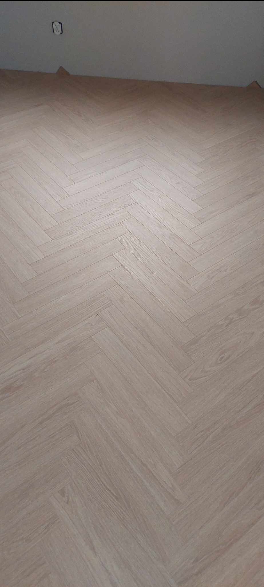 Jasne panele podłogowe ułożone w jodełkę, widoczny fragment ściany z gniazdkiem elektrycznym.