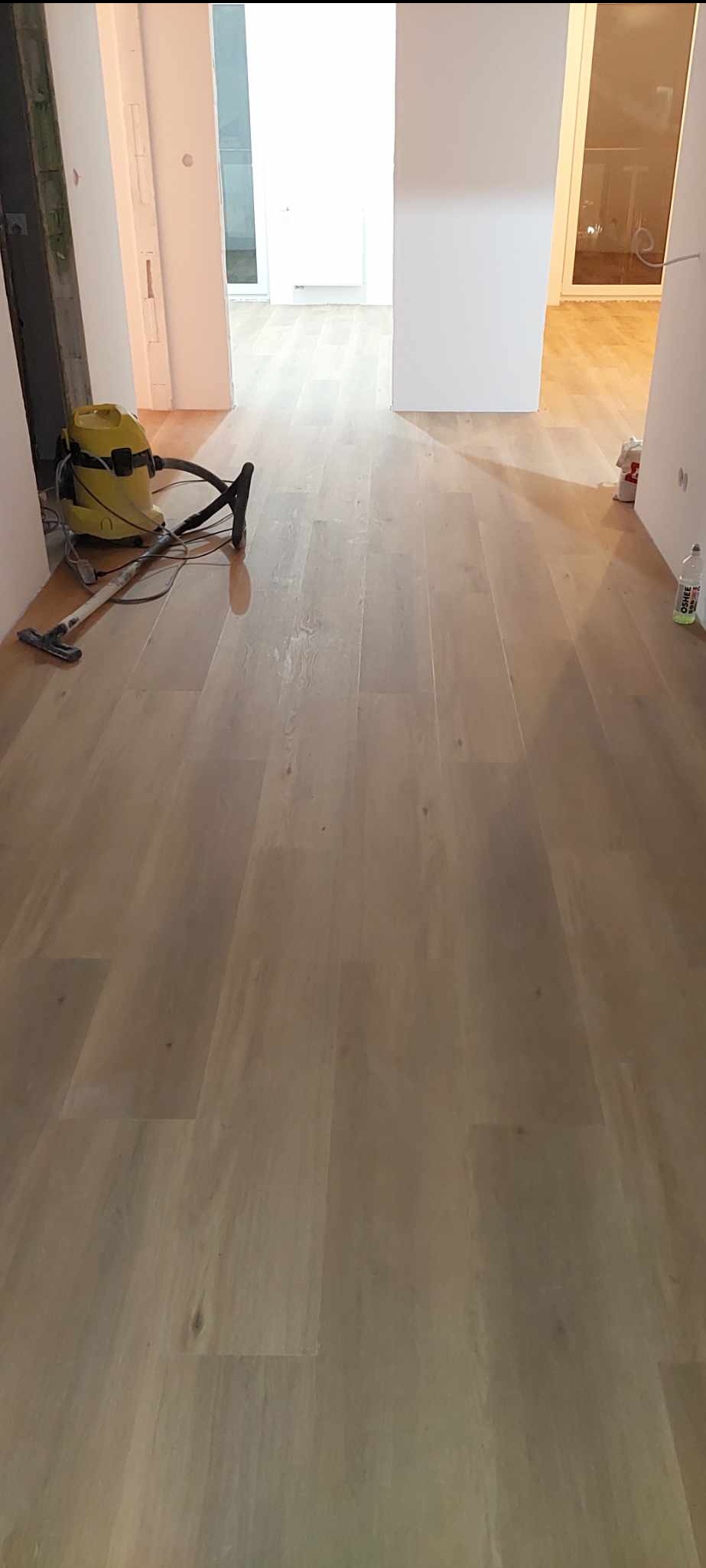 Jasna, drewniana podłoga z widocznym odkurzaczem przemysłowym i butelką płynu do czyszczenia, w tle widoczne kolejne pomieszczenia.