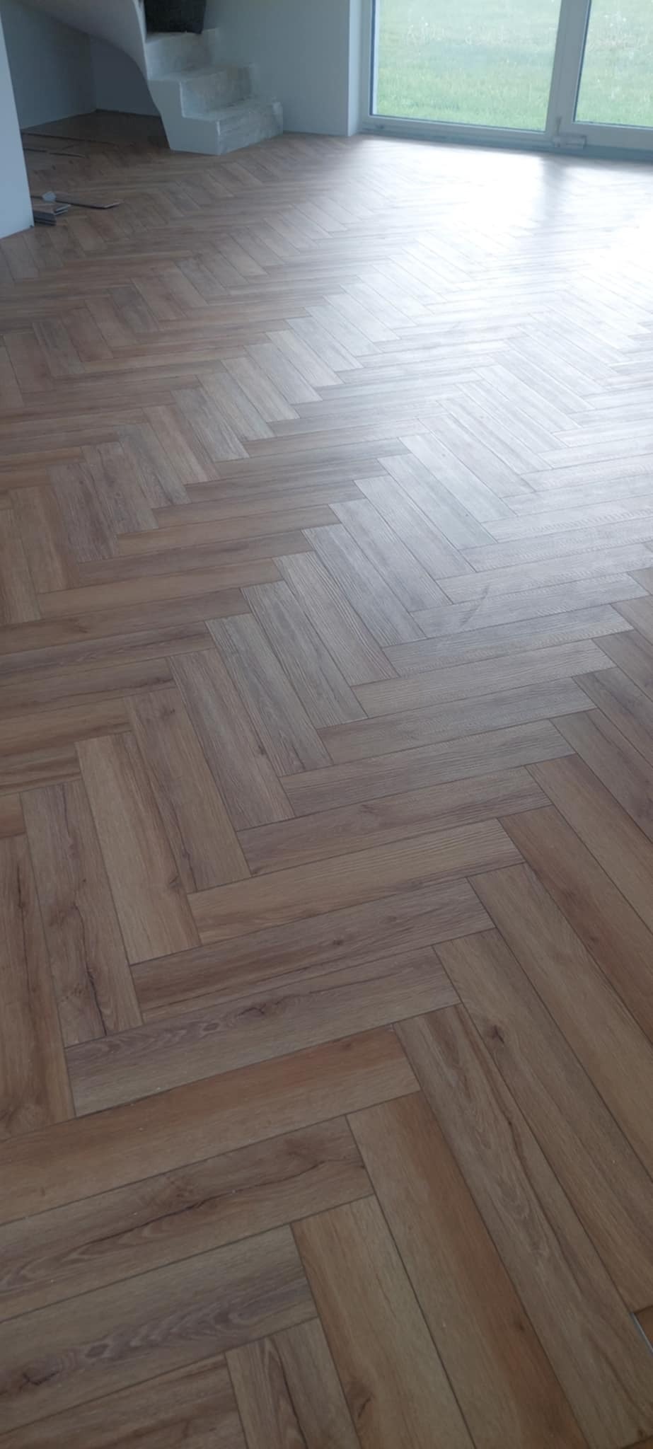 Ułożona podłoga z paneli w jodełkę, widok na schody i okno. Jasne wnętrze po remoncie, naturalne odcienie drewna. Wykończona podłoga w nowym domu.