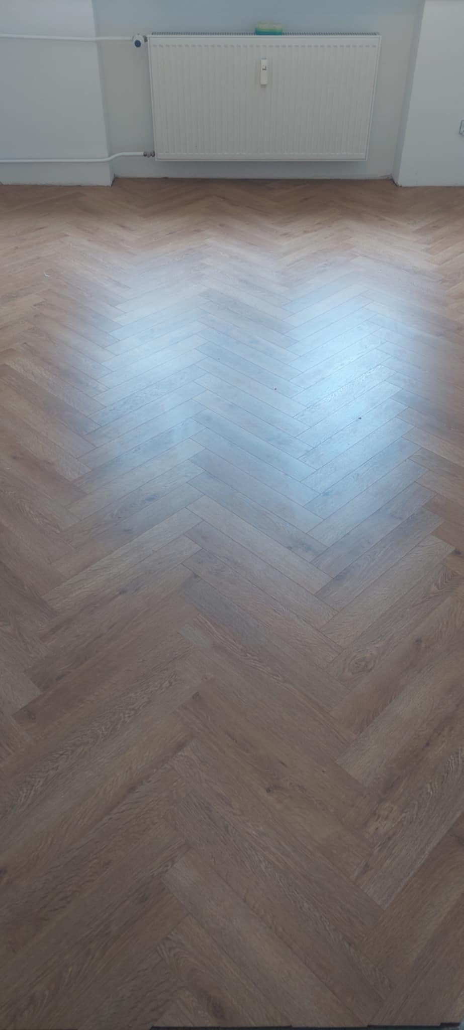 Ułożone panele podłogowe w jodełkę, brązowe, z widocznym grzejnikiem na ścianie. Widok z góry, równomierne oświetlenie.