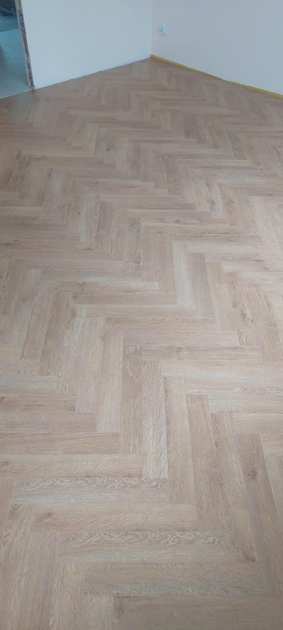 Jasne panele ułożone w jodełkę na podłodze w nowym pomieszczeniu, widok z góry. Ściany pomalowane na biało, widoczne listwy przypodłogowe.