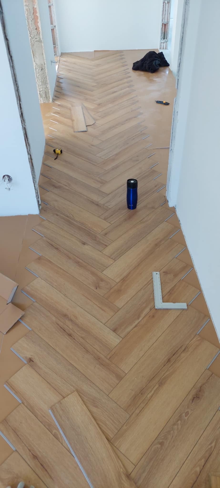 Układanie paneli w jodełkę, widoczne narzędzia: miarka kątowa, poziomica, niebieski bidon. Częściowo ułożona podłoga, brązowe panele z widoczną strukturą drewna.